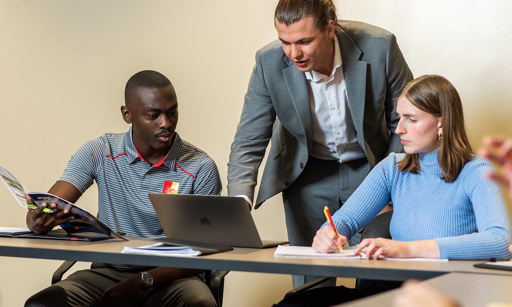 A teacher standing and two students sitting looking at a laptop in the Kelce College of Business