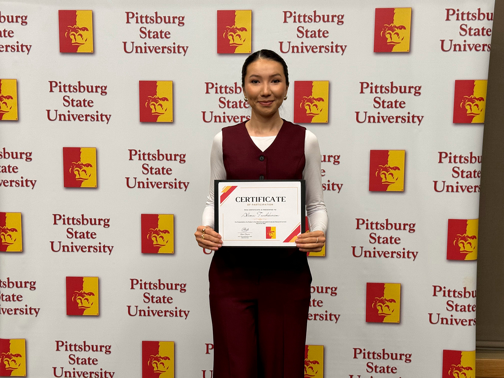 Student poses with certificate infront of pitt state backdrop
