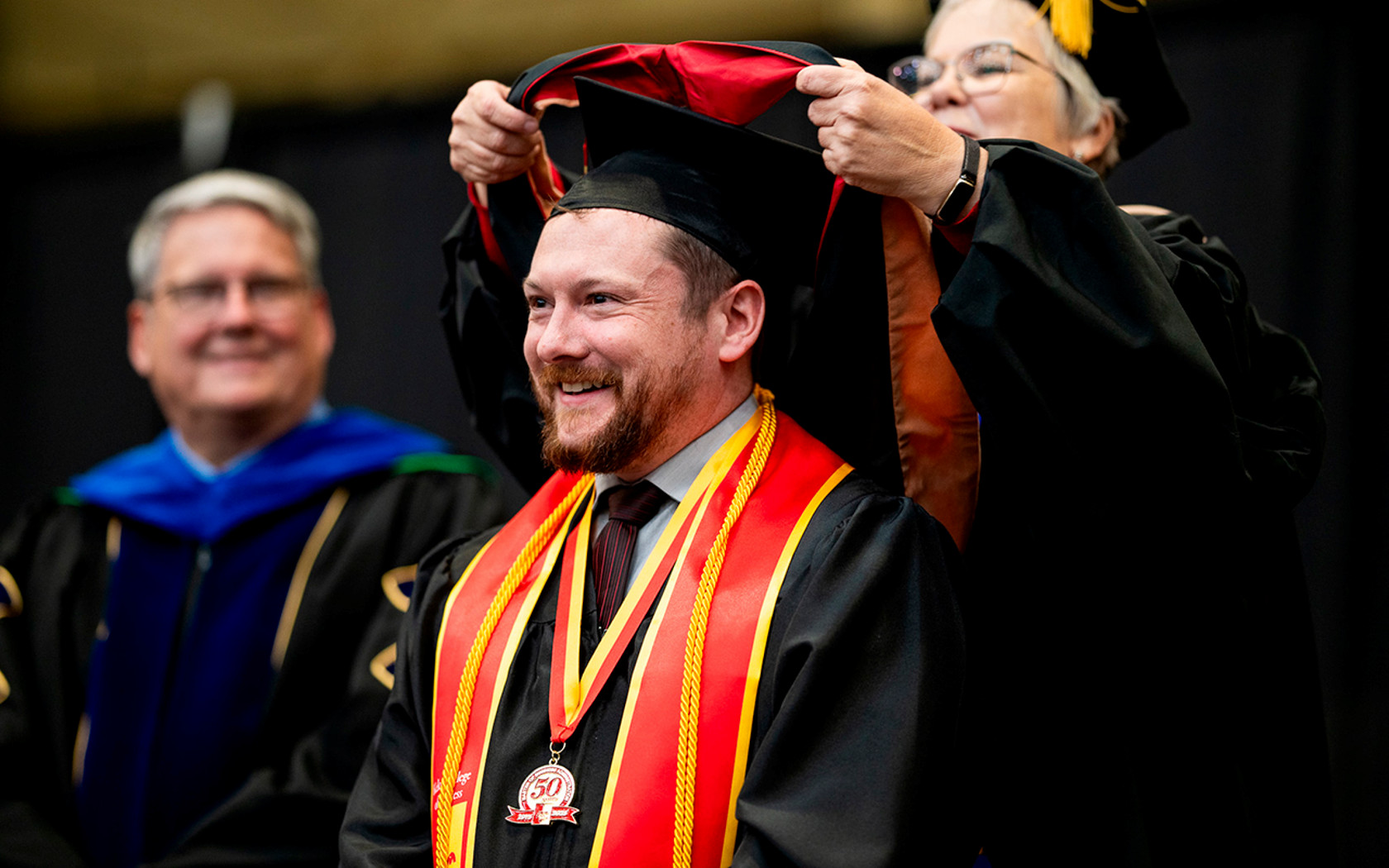 Man sits in graduation gear and gets his stole/scarf put on at the weeds, smiling ear to ear