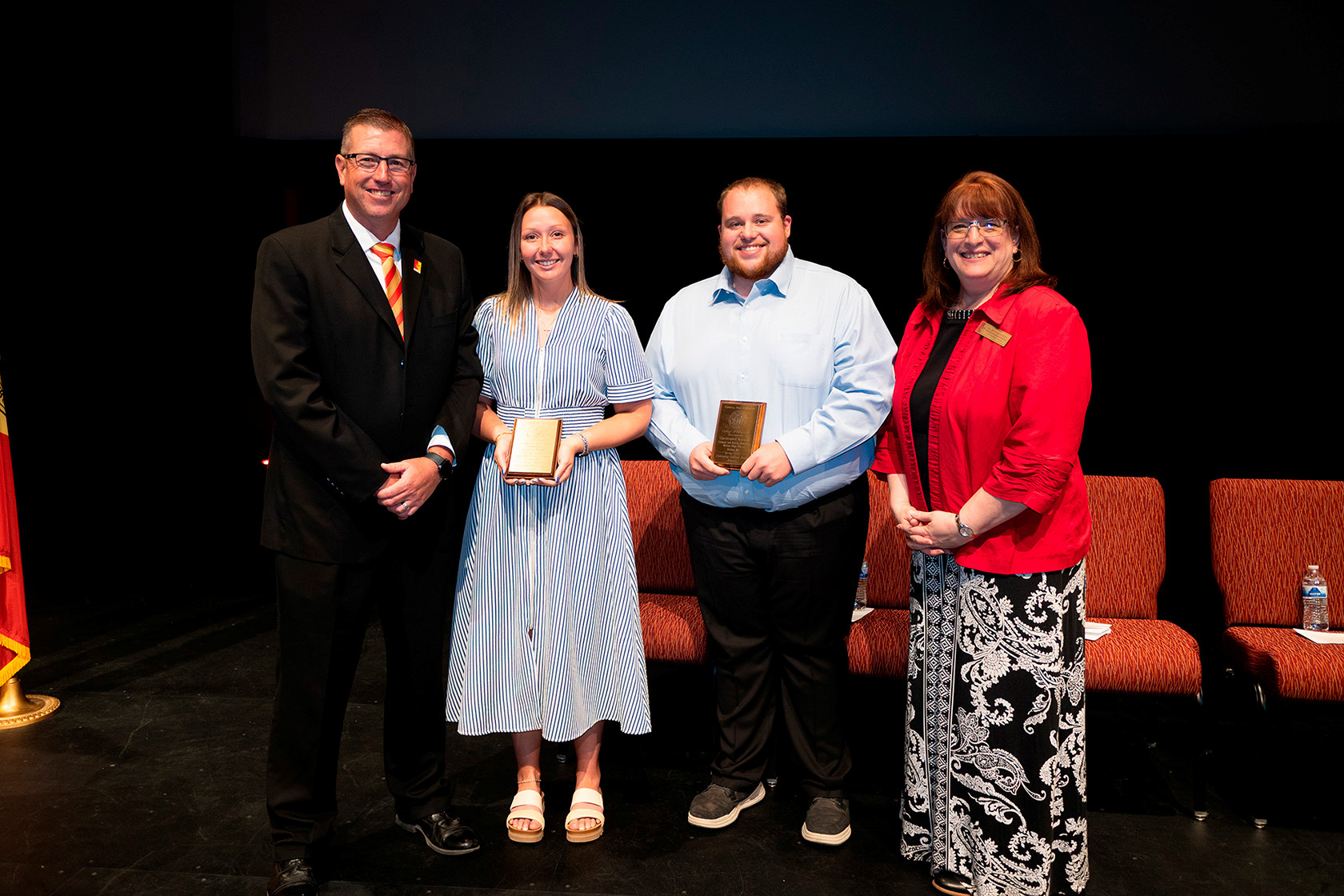 Four people on stage; two in the center hold award plaques