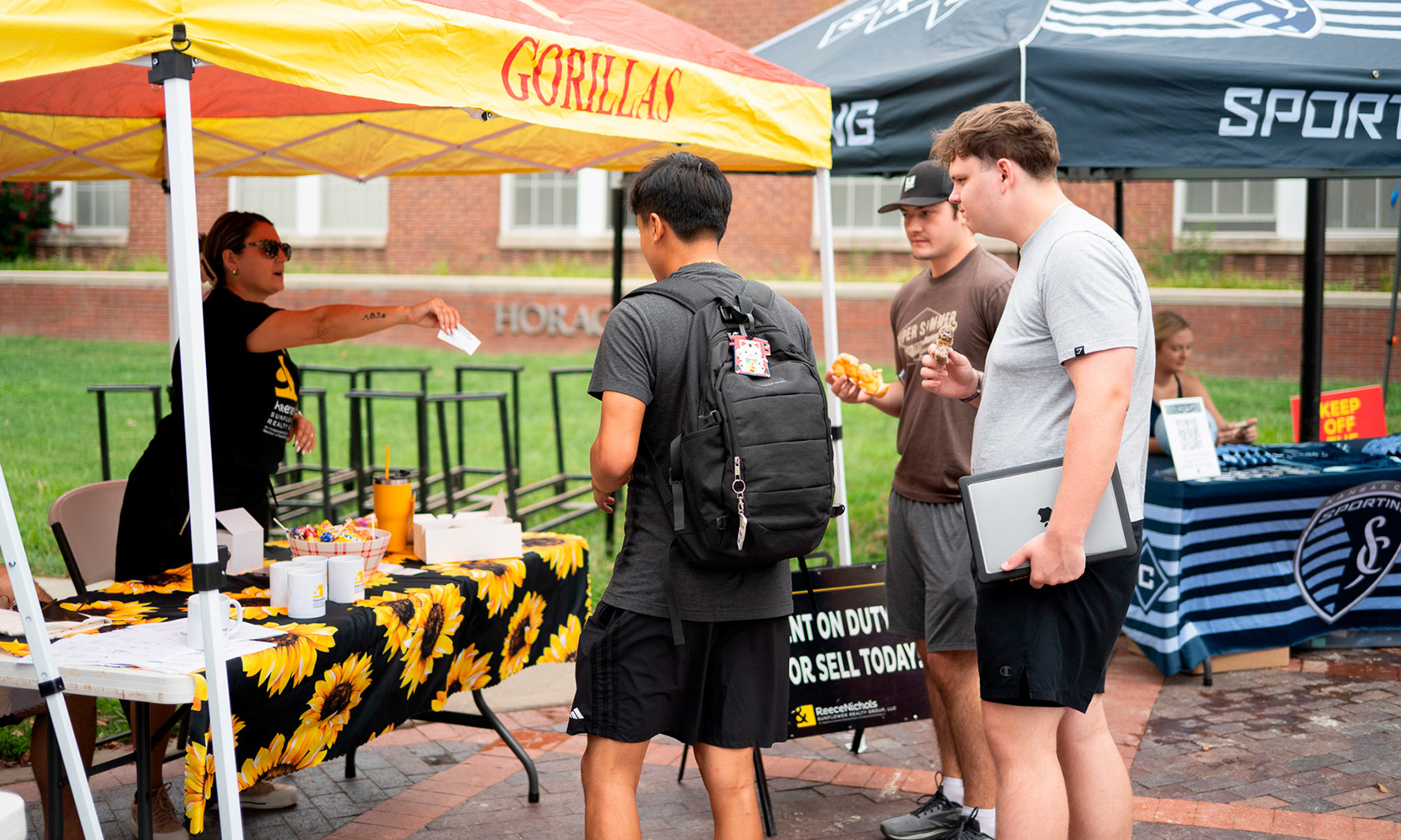 three male college students interacting with female realtor at booth during outdoor community fair