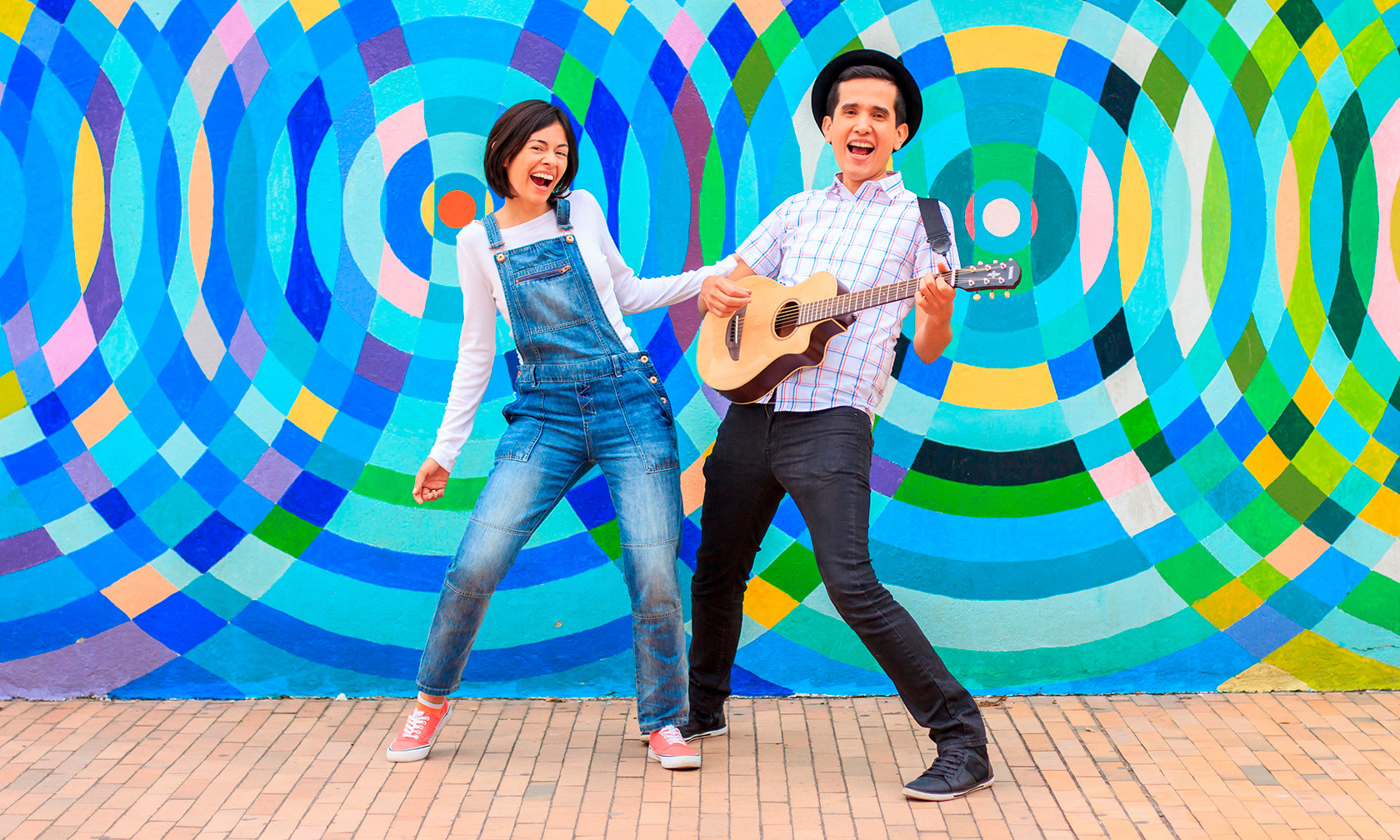 a woman and man smiling in front of a very colorful background, the man has a guitar