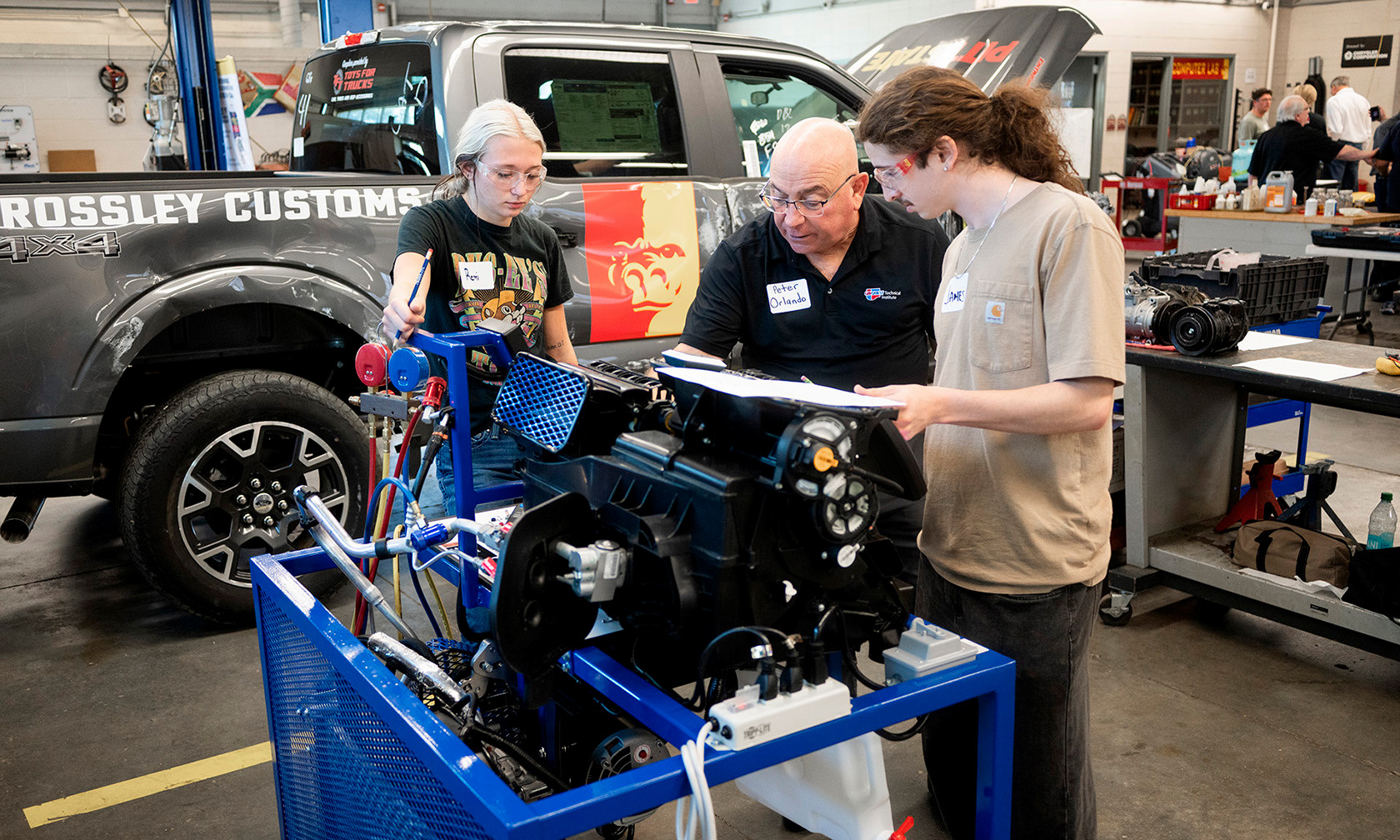 An instructor reviews a document with two students beside a car engine on a stand in an automotive workshop.