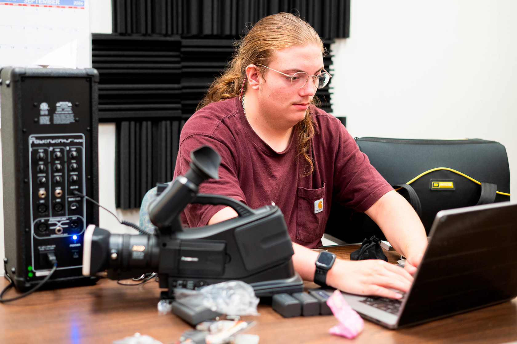 A student works at a laptop in a media production studio, with a video camera and audio mixer on the table and acoustic foam panels on the wall behind them.