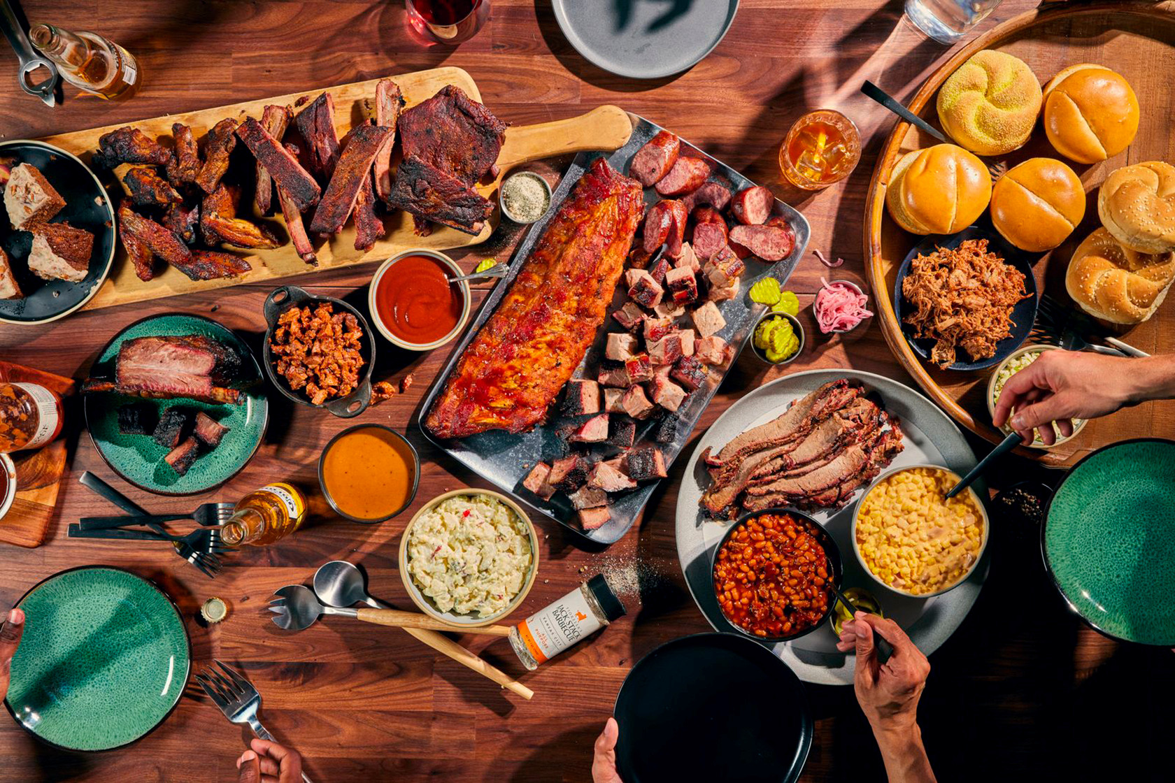 On a gray table sits a variety of bbq foods and sides, including brisket, ribs, pulled pork, and sausage