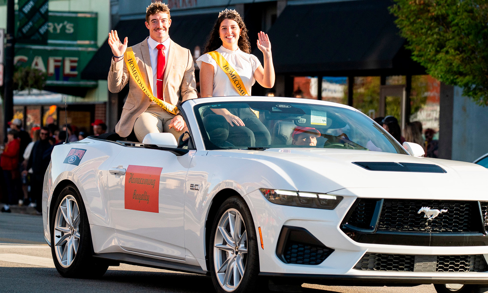 two students ride in a car in the 2025 Homecoming parade. They are wearing sashes and crowns and are waving.