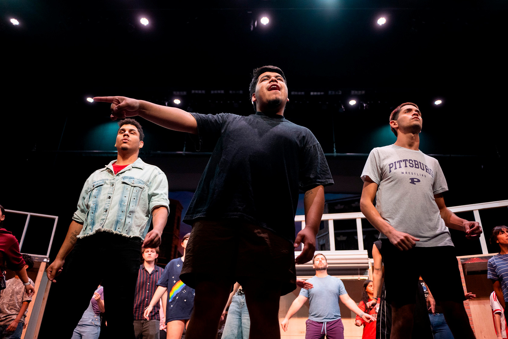 Three male students stand at the front of the stage; the center student points and sings upward while the full ensemble performs behind them.