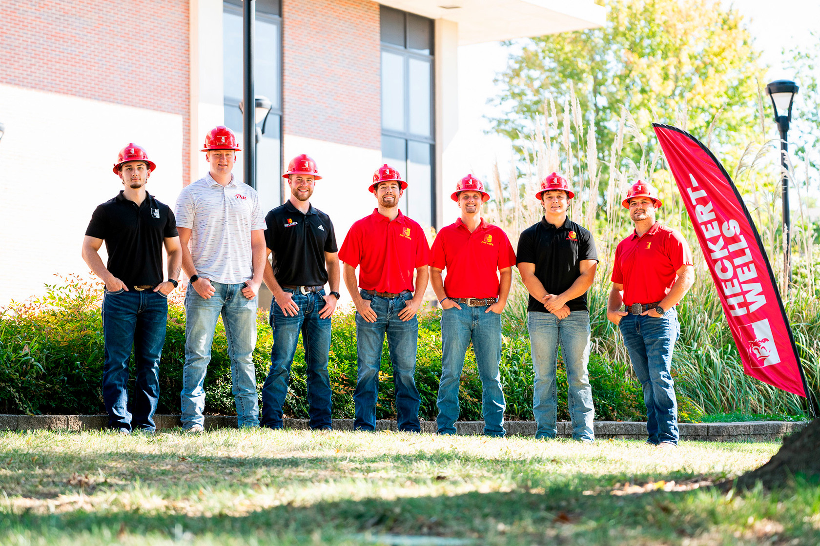 A group of seven students wearing hard hats standing outside of a campus building