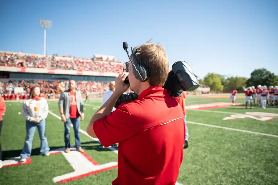 a student uses a video camera to record the game and halftime show on the field