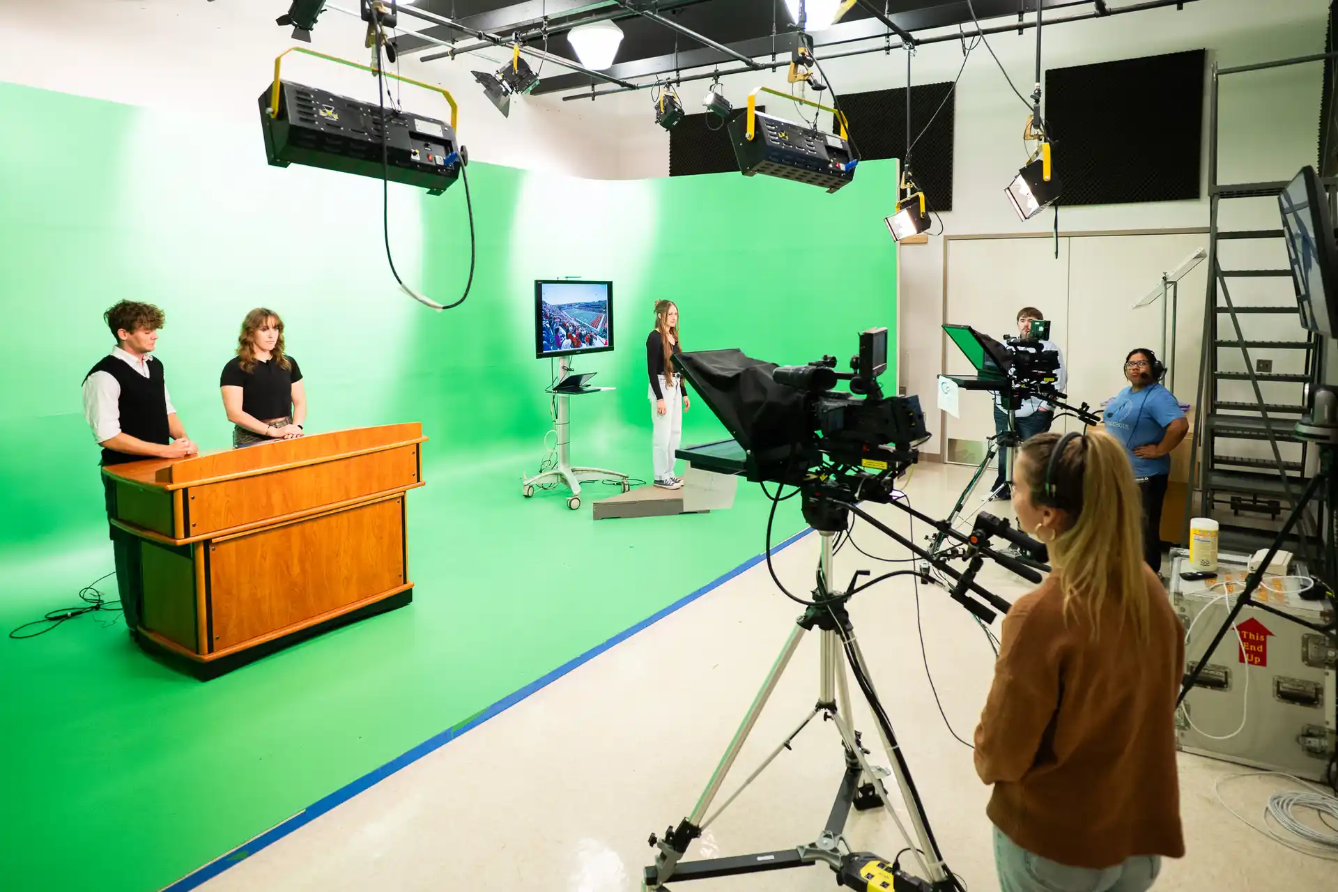 Students stand on set and behind camera infront of a green screen 