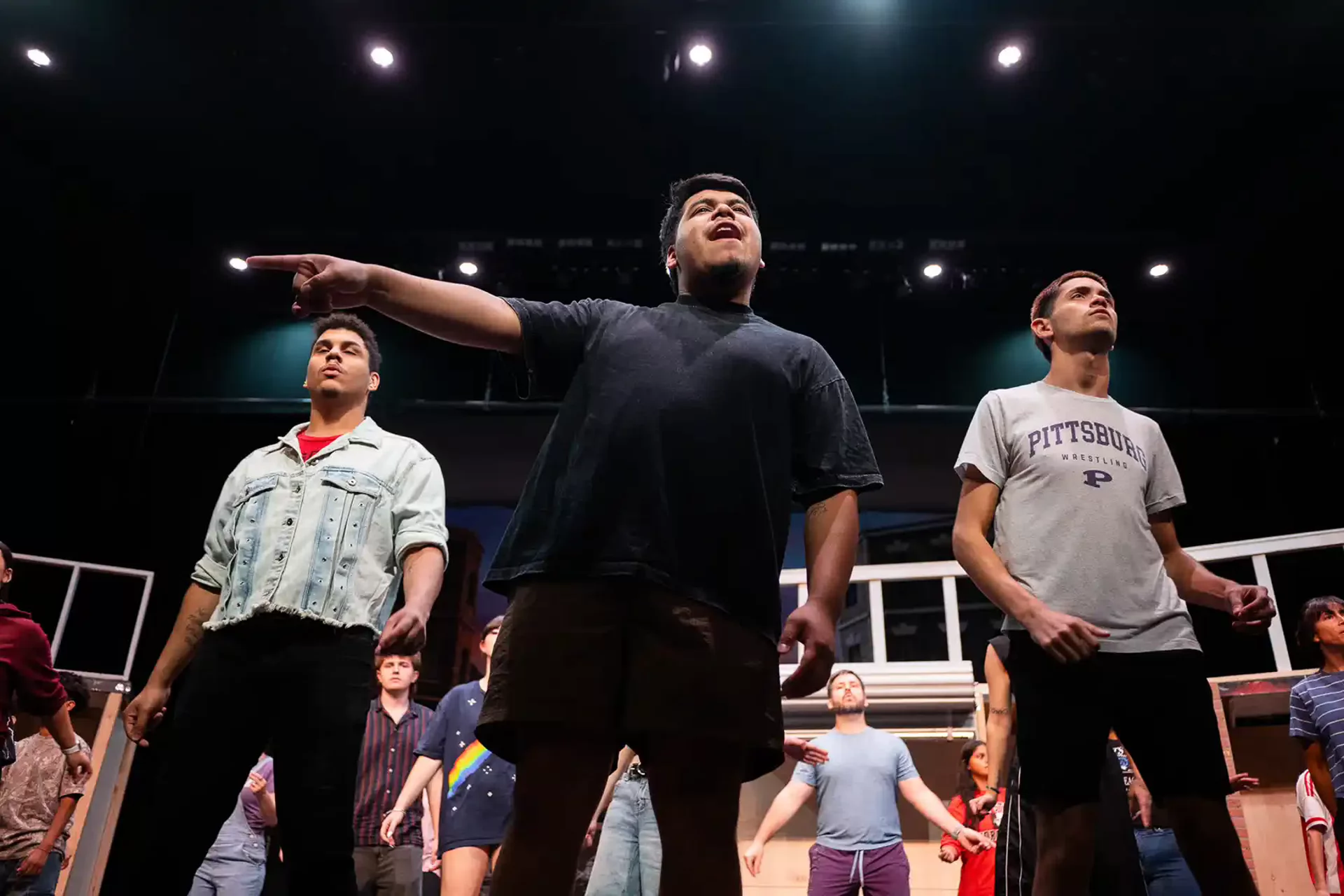 Three male students stand at the front of the stage; the center student points and sings upward while the full ensemble performs behind them.