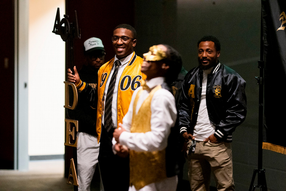 Alpha Phi Alpha fraternity members in coordinated black-and-gold jackets stand at a new member presentation, smiling and watching as a performer in a gold vest stands in the foreground